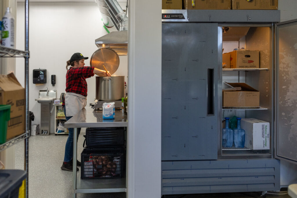 Preparing food in a commercial kitchen, a woman stirs a large pot os steaming soup while a commercial refrigerator keeps the produce cool.
