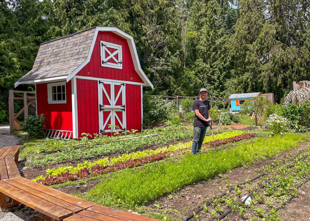 A man stands in the One Tiny Farm garden, working with garden tools, in front of a red barn.