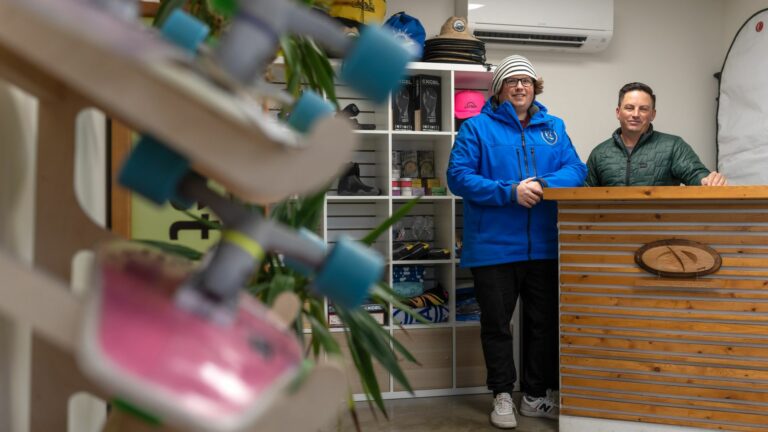 Compass Adventures owners pose in their surf shop, which sells long boards, surf boards, wax, and other essentials for time on the water.